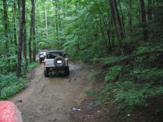 Two 4x4 SUVs driving on a muddy trail through a dense forest. The lead vehicle is a 1978 Ford Bronco