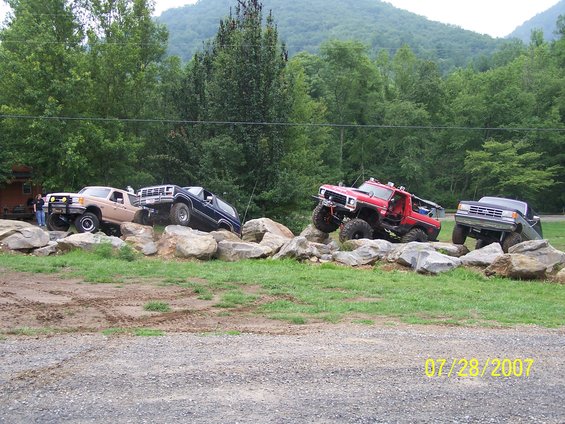 Four modified trucks navigating a rock obstacle course; tan, blue, red, and gray vehicles are visible