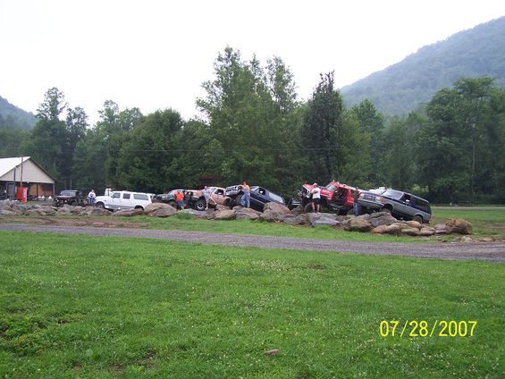 Several modified trucks and SUVs are positioned on large rocks, with people standing around them