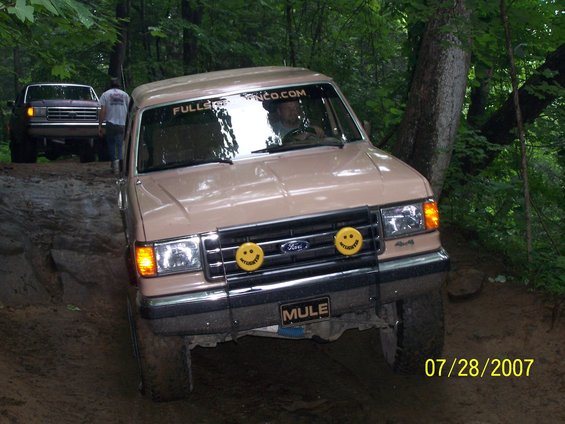 Tan Ford F-Series truck with smiley face lights and 'MULE' plate, another truck behind it on a dirt trail