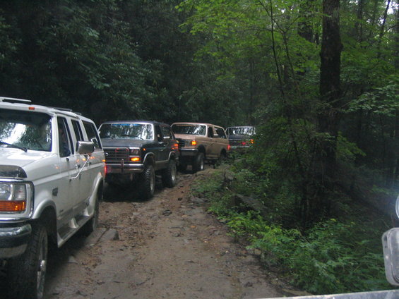 Four SUVs, including a white Ford Econoline van and three Ford Broncos, drive in a line on a muddy forest road