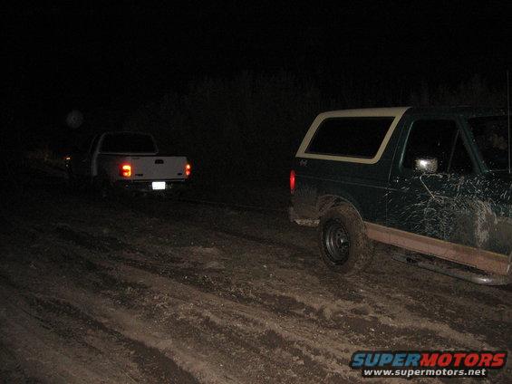 green Bronco and a white pickup truck are on a muddy dirt road at night. The Bronco is in the foreground