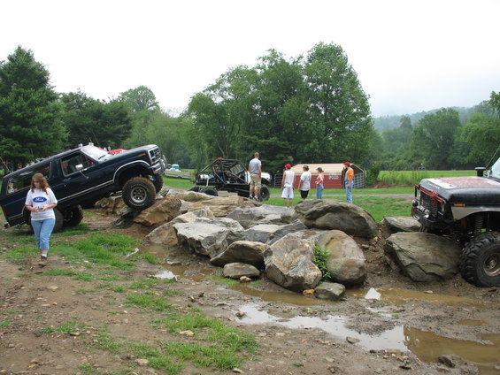 Off-road vehicles on a rocky course with people watching; one vehicle is a dark SUV, another is a buggy, and a third is a red-accented truck