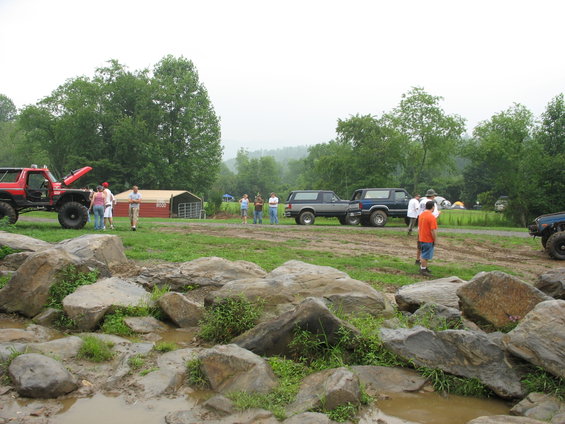 Several off-road vehicles, including a red Jeep-like vehicle with hood up and two blue SUVs, parked in a muddy, rocky area with people standing around