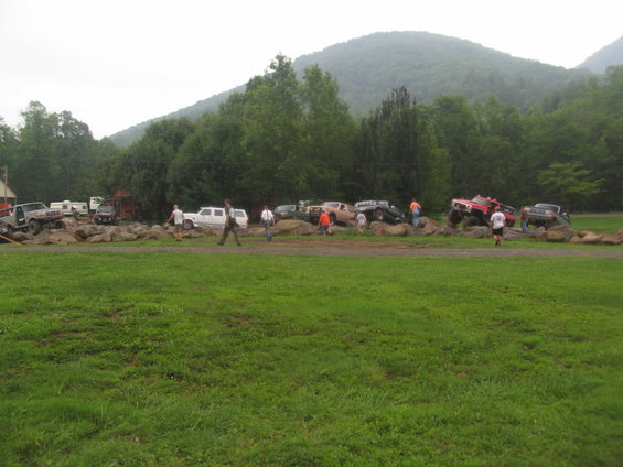 Several off-road vehicles are positioned on a rocky obstacle course with people standing around. Mountains and trees are in the background
