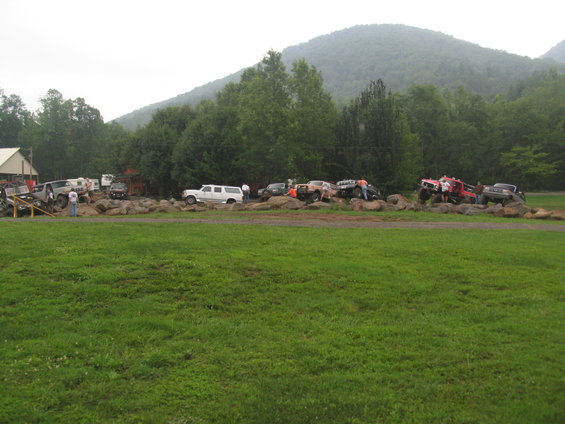 Several off-road trucks and SUVs are positioned on a rocky terrain with a wooded hillside in the background