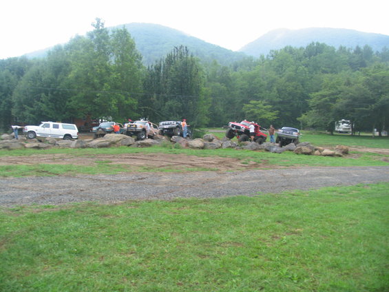 Multiple off-road vehicles maneuvering over a rocky obstacle course with trees and mountains in the background