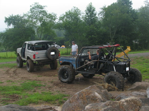 Two off-road vehicles, one a modified buggy and the other a lifted SUV, are on a muddy trail with trees in the background