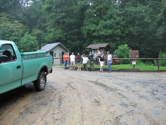 Green pickup truck, side view, parked on a muddy road near a group of people and buildings