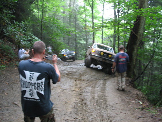 Brown Ford F-Series truck and a dark SUV on a muddy forest trail, with people taking photos