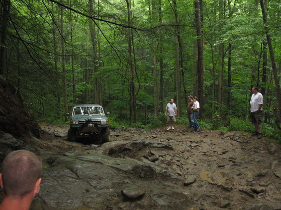 Green off-road vehicle navigating a muddy, rocky forest trail with people watching