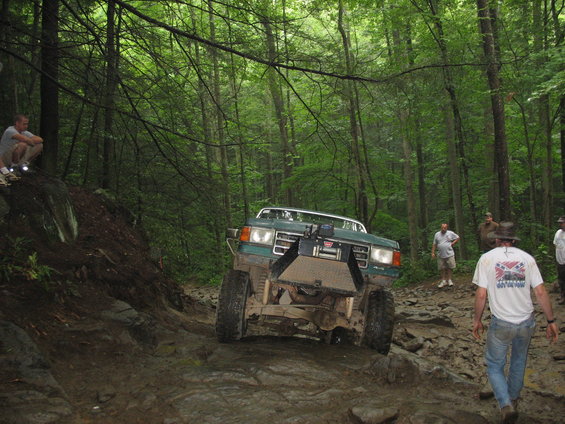 Green off-road truck navigating a steep, rocky path in a dense forest. People watch from the side