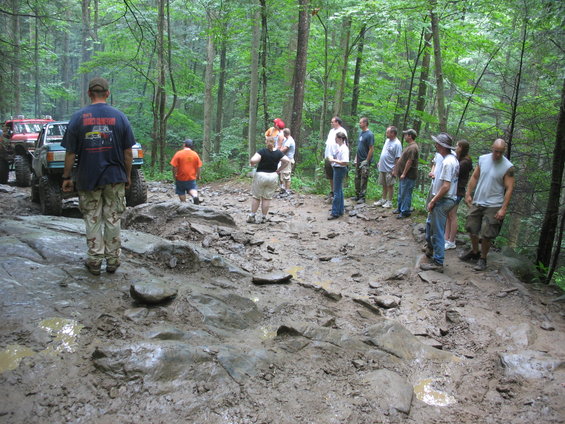 Group of people watching off-road vehicles navigate a very muddy and rocky trail in a forest
