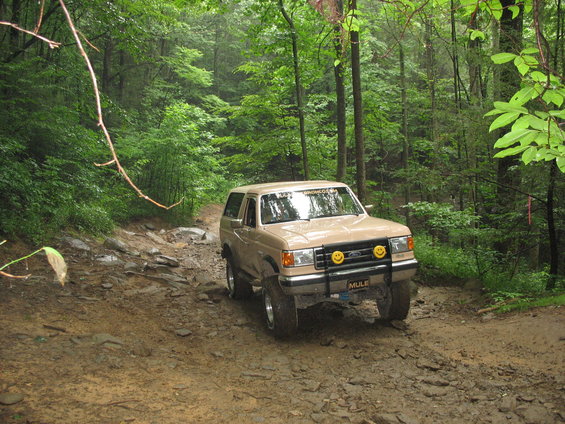 Tan Ford Bronco off-roading on a muddy trail, front 3/4 view. Visible text 'BRONCO.COM' on windshield and 'MULE' on bumper