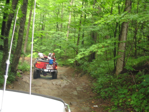 Red pickup truck with people in the bed driving on a rocky forest trail. Another vehicle is partially visible in the foreground