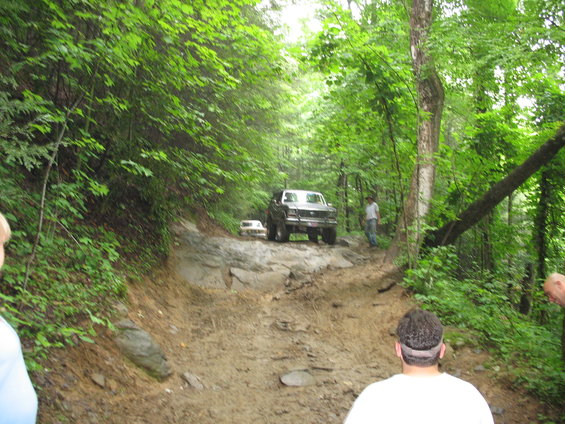 Two trucks driving up a rocky, muddy trail in a forest