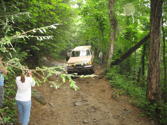 Two off-road vehicles navigate a muddy, wooded trail; one vehicle is in the foreground