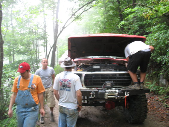 People gathered around a red pickup truck with its hood up on a dirt road