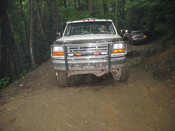 Front view of a white Ford F-Series truck on a muddy trail. Another truck is visible behind it