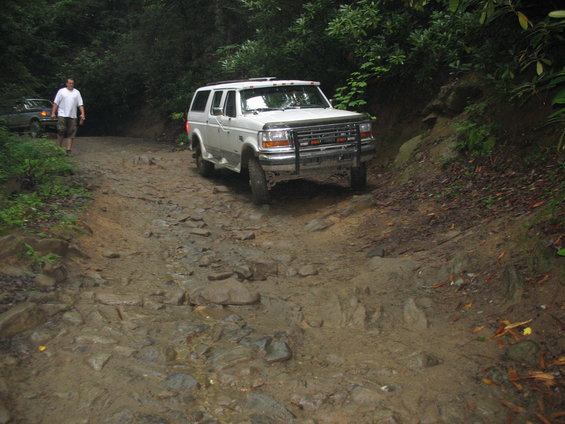 White SUV driving on a rough, muddy, and rocky trail in a wooded area. Another vehicle and a person are visible in the background