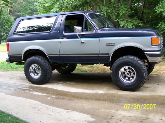 Two-tone blue and gray Ford Bronco with chrome wheels and off-road tires, parked on a wet driveway