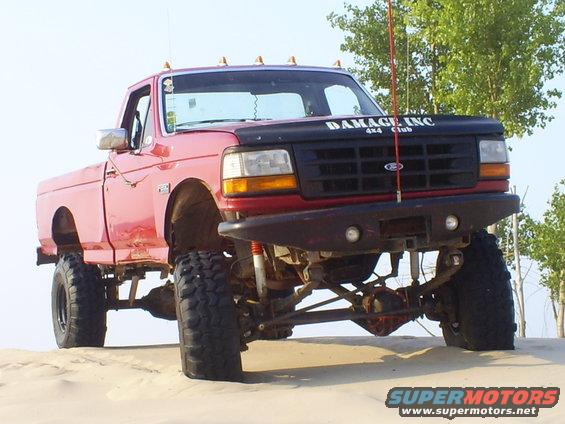 Lifted red pickup truck with large tires, custom bumper, and "DAMAGE INC 4x4 Club" on the hood