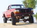 1994 Ford F-250 - A heavily modified red pickup truck sits on sand dunes under a clear sky.