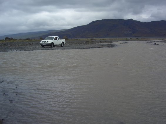 White pickup truck parked on a rocky shore next to a wide, murky river under a cloudy sky
