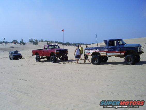 Two lifted pickup trucks and a small SUV parked in sand dunes under a clear blue sky. One truck is red, the other is blue and white with red stripes