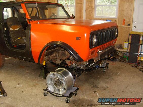 Orange International Harvester Scout II with black fenders, missing front wheel, on jack stands in a garage
