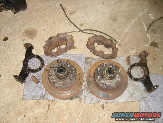 Two brake rotors with hubs, two brake calipers, and two steering knuckles laid out on a concrete floor