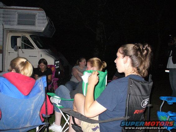 People gathered around a campfire at night, with an RV parked nearby