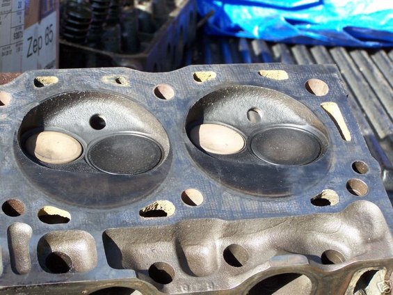 Close-up of an engine cylinder head with two sets of intake and exhaust valves visible. Some debris is present around the ports