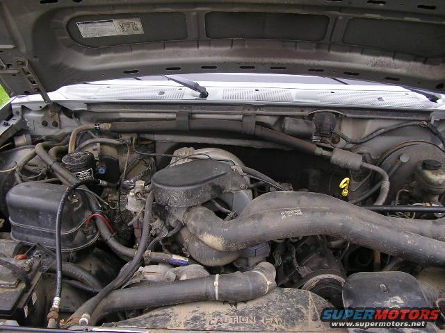 Engine bay with hoses, wires, and engine components visible. Text on a hose reads 'CAUTION FAN'