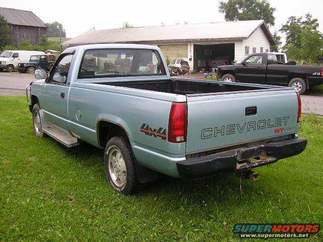 Light blue Chevrolet pickup truck parked on grass, rear view. Another black pickup is parked in front of a garage