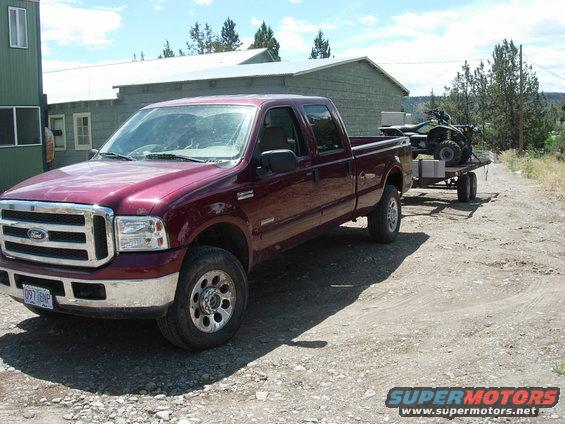 Maroon 2005 Ford F-350 Super Duty pickup truck, crew cab, long bed, towing a flatbed trailer with an ATV on it. Parked on a gravel driveway in front of a green building
