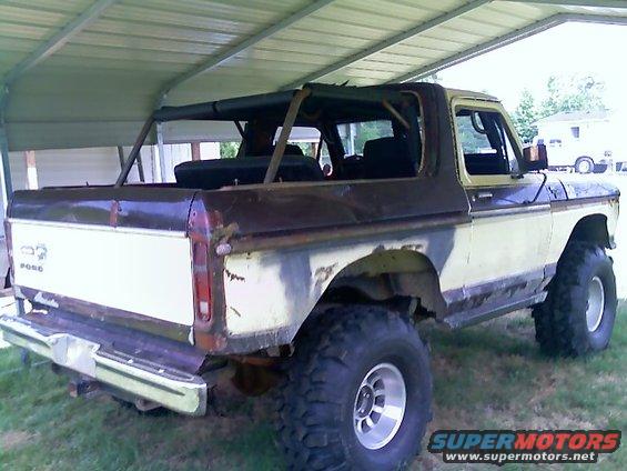 Rear 3/4 view of a lifted, two-tone Ford Bronco with a soft top removed. It has large off-road tires and appears to be in rough condition