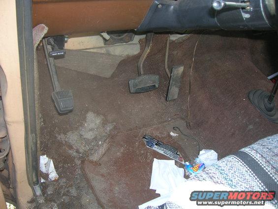 Vehicle interior floor with three pedals, brown carpet, and debris. A Hot Wheels package is visible