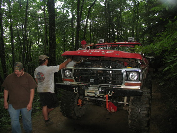 Red off-road truck with hood open, winch, and off-road tires. Three men are visible