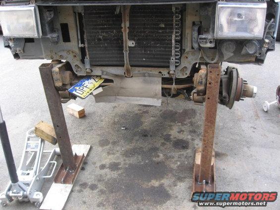 Front end of a truck, missing its hood and fenders, is supported by jack stands. A license plate is visible