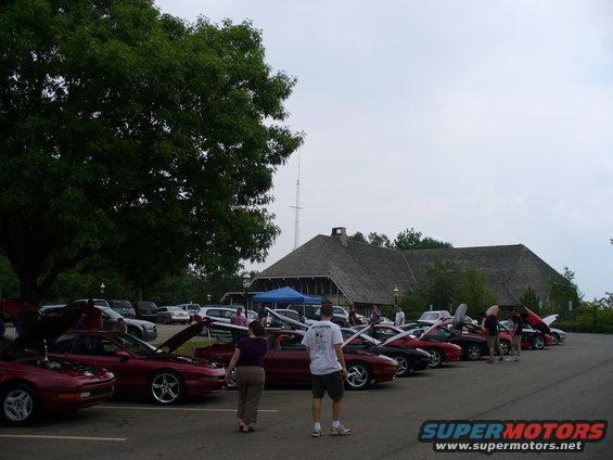 Several red cars parked in a lot with hoods up, people milling around. Building in background