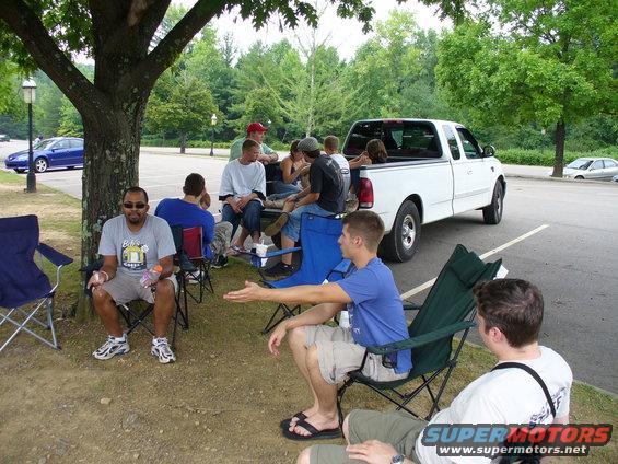 White pickup truck parked in a lot with people sitting in chairs nearby. A blue car is visible in the background
