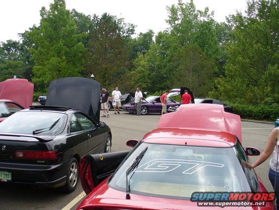 Several cars with hoods open at an outdoor gathering. A black coupe is in the foreground left, a red coupe is in the foreground right
