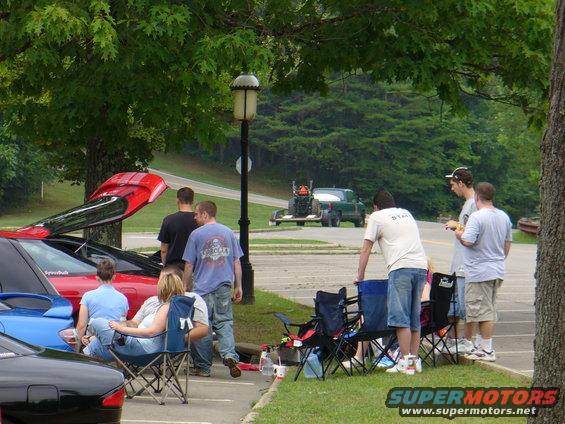 Red car with trunk open, parked next to a blue car and a black car, with people standing and sitting around