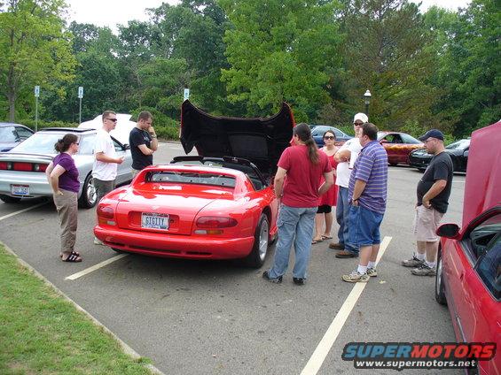 Red sports car with hood open, license plate reads GIGITY. Several people are looking at it. Another silver car is parked to the left