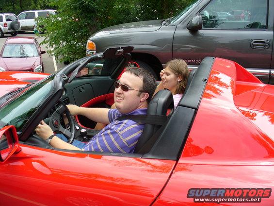 Red convertible sports car with a man driving and a woman in the passenger seat. A grey truck is parked behind