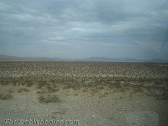 Desert landscape with scrub brush under a cloudy sky, with mountains in the distance. TheWebWheeler.com watermark is visible