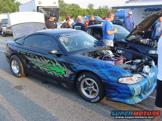 Black Ford Mustang with blue and green flame graphics, hood open showing engine. Another Mustang is parked next to it
