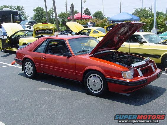 Orange 1986 Ford Mustang coupe with hood open, parked at a car show