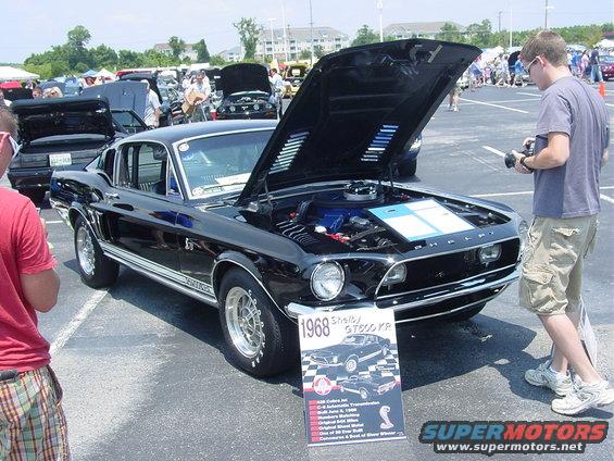 Black 1968 Shelby GT500 KR Mustang with hood open, a sign in front, and people around
